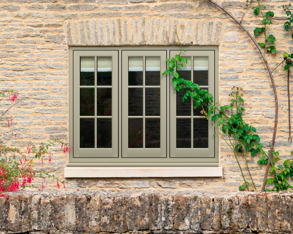 Sage green triple-pane windows on a stone cottage wall surrounded by climbing plants and flowers
